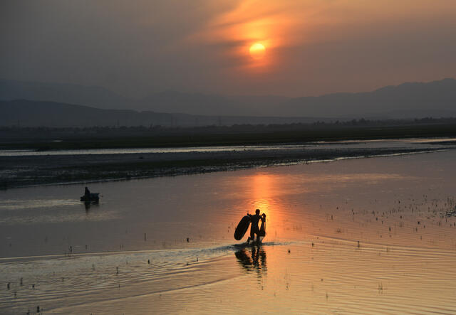 A sunset photo on a beach in Afghanistan. There are mountains are in the background and the sillhouettes of two people with small boats and fishing nets, one in the water and the other walking into the water.