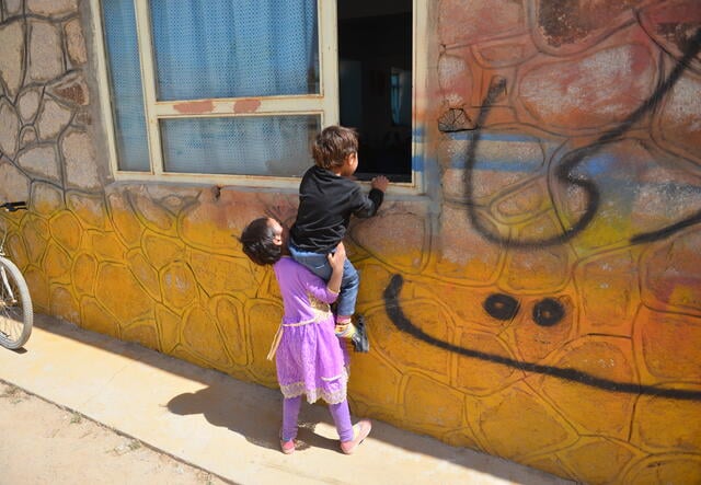 A young girl picks up a young boy to look in the window of a colorful building 
