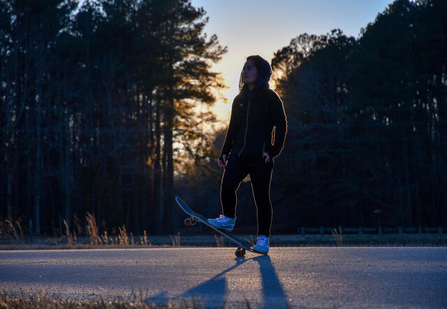 In the evening and in front of a forest, Belqisa stands on her skateboard and looks into the distance. 