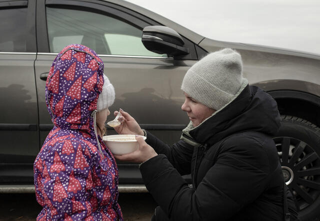 A mother kneels down to feed her daughter, both are outside wearing winter clothes 