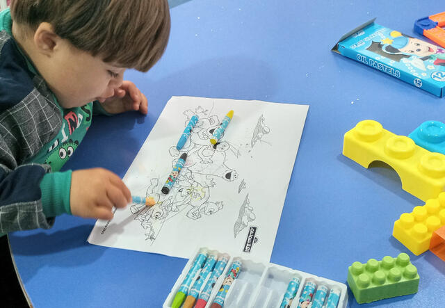 Five-year-old Syrian boy Saeed sits leaning over a low table, using crayons to color in a page with Sesame Street characters.