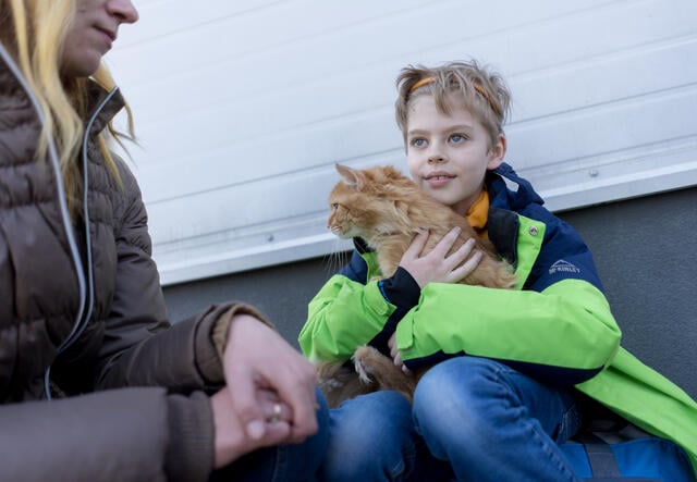 A young boy wearing a headband and coat sits against a wall, holding an orange cat.