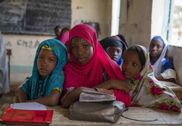 Three girls sit with books and look at the camera, with other children sitting behind them.