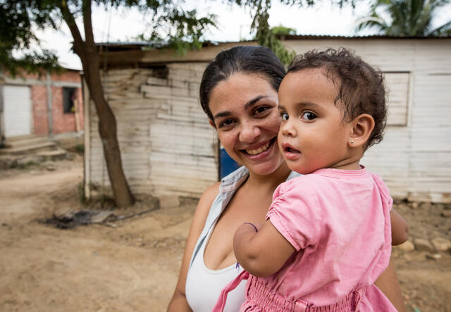 A woman smiles and holds her daughter, who is wearing a pink dress.