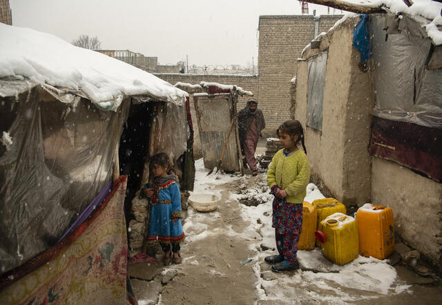 Two young girls stand outside of a tent in the snow with a man walking behind them.