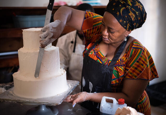 A woman spreads white frosting on a three-tiered cake.