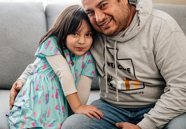 A father and daughter sitting on a couch and smiling in their new home they bought with the assistance of the International Rescue Committee's IDA savings program. 