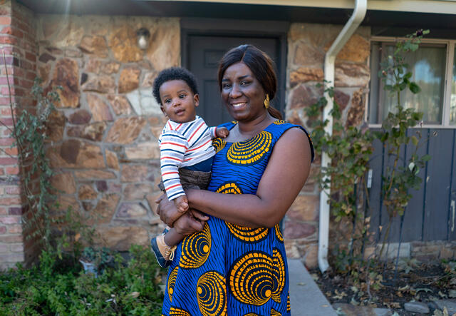 A woman in a blue and yellow dress holds her one year old son and smiles while standing in front of a house 