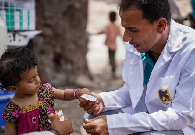 A seated male nutritionist gives a young girl a packet of vitamin-packed peanut paste for malnutrition.