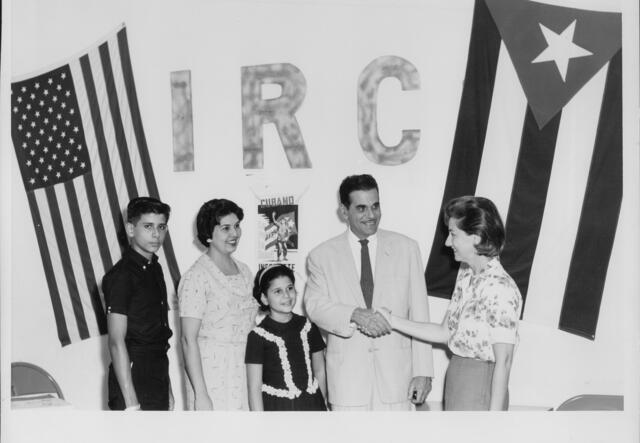 A Cuban family of four poses with an IRC staff member in front of a wall that says "IRC," next to a Cuban flag and a U.S. flag.