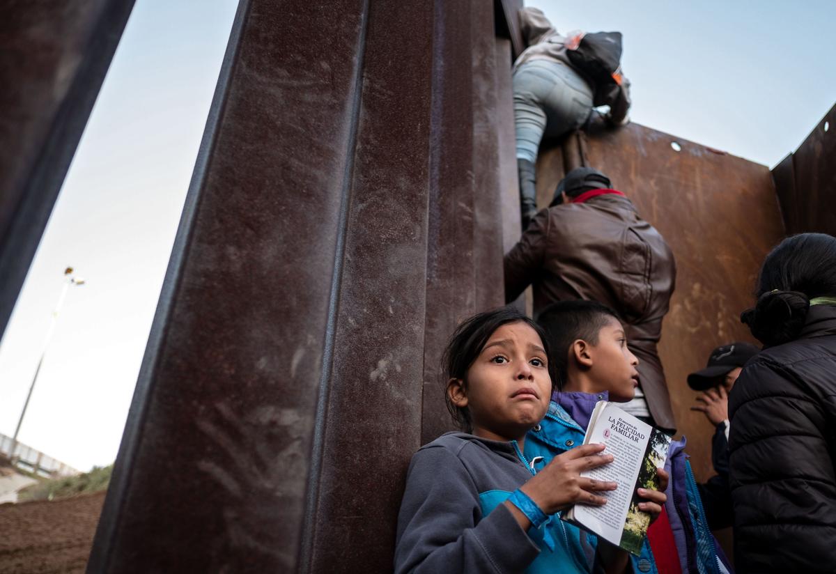 A Central American girl holds a book as others traveling in a caravan climb the Mexico-U.S. border fence in an attempt to cross to San Diego County.