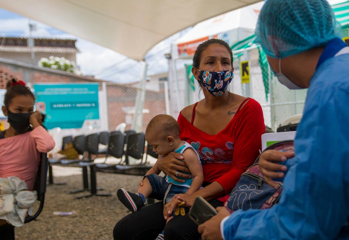 A health worker in protective equipment speaks with a woman who is sitting wearing a mask and holding her baby