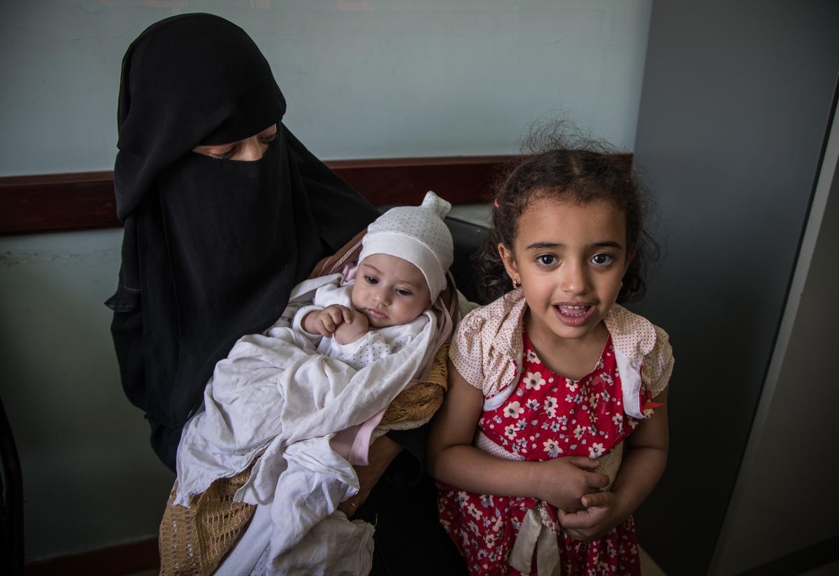 Mona holds her baby while her older daughter stands nearby.