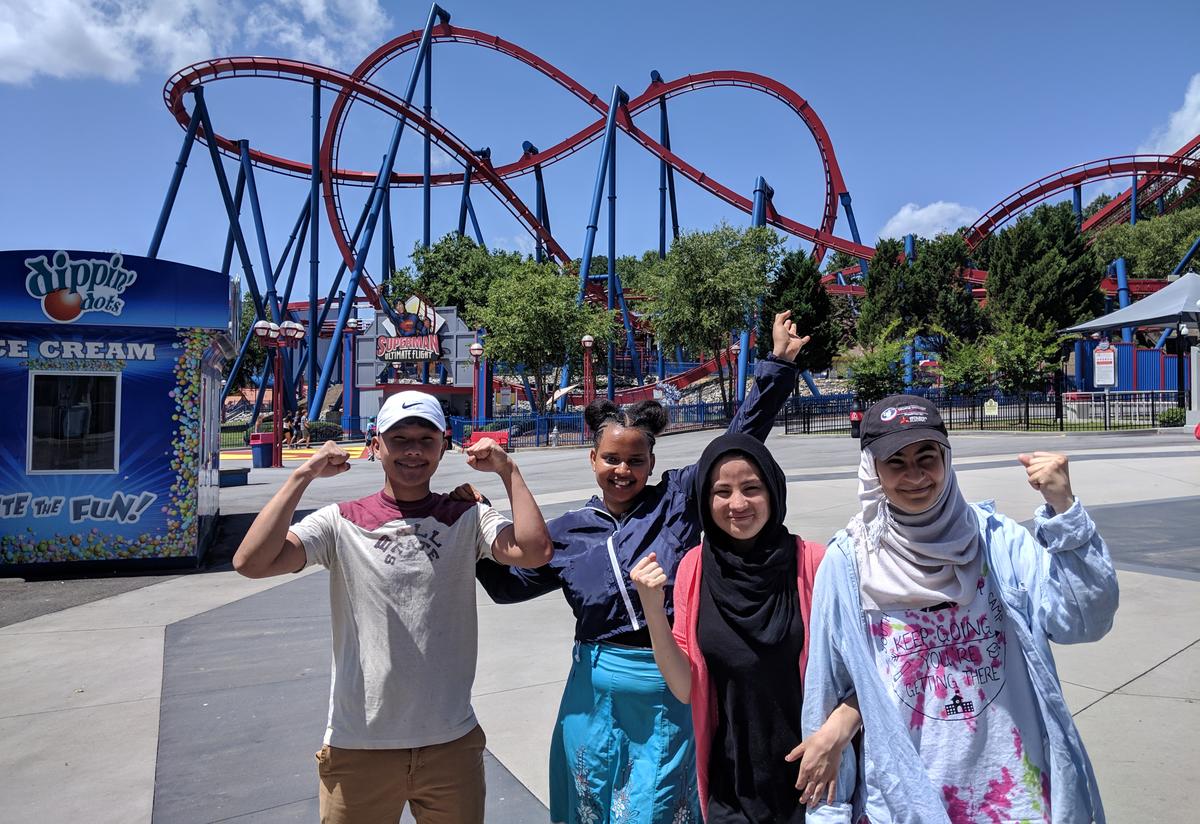 IRC Youth Futures volunteer, Elise Petersen, raised money to treat all of our campers to a fun-packed day at Six Flags amusement park. Four students post for a picture in front of a large rollercoaster.