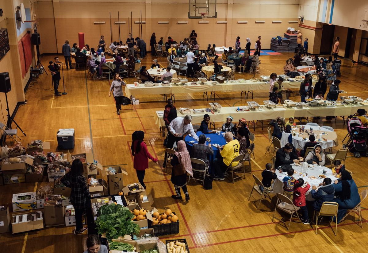 The IRC in Atlanta's 18th annual Thanksgiving Feast. A large church gym hall is filled with circular tables, each with diverse groups of families, volunteers and IRC staff eating together. Two large long banquet tables in the center of the room are filled with Thanksgiving food as people serve themselves.