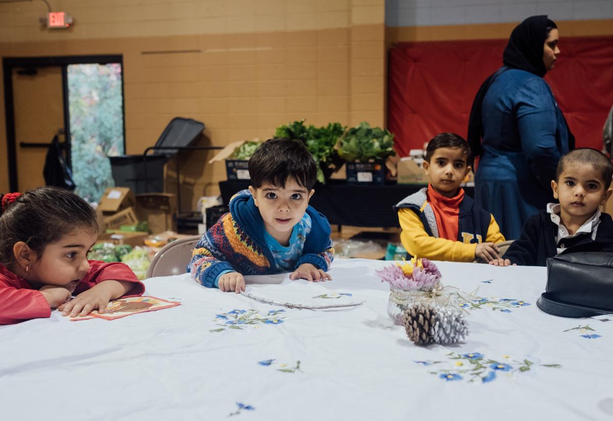 Four small children under five years old sit around a circular table waiting for their meal. One boy in the center of the photo is leaning across the table and gazing directly into the camera with big brown eyes.
