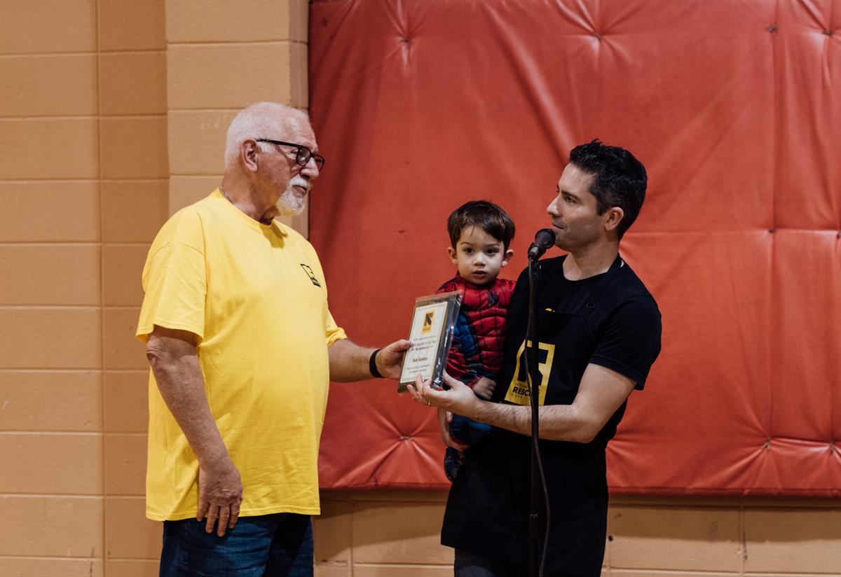 The IRC in Atlanta's 18th annual Thanksgiving Feast. The IRC's volunteer coordinator, Duncan, presents the volunteer of the year award to volunteer Bob Gordon, an older gentleman wearing an IRC t-shirt. Duncan is holding his young son who is dressed as Spiderman.