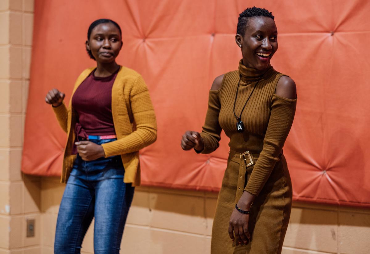 Two young women from the IRC's Youth programs dance, side by side as part of the entertainment portion of the feast. They are wearing brightly colored knitwear and are smiling and laughing together.