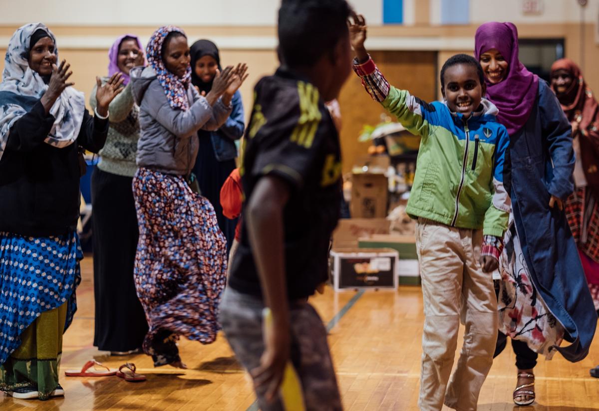 A group of women in colorful hijabs jump in the air, smile and clap their hands to music. Two young boys dance in the center of the photo. The end of the feast turned into a group dance party!