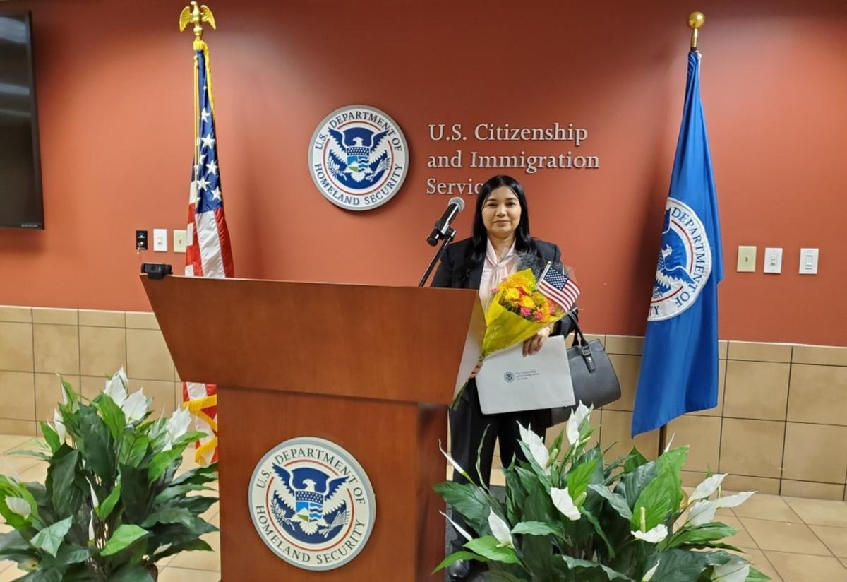 Safa at USCIS, just after being sworn in as a new American citizen. A short woman with dark hair stands behind a podium holding a bouquet of flowers and a paper certificate. The words "United States Citizenship and Immigration Services" are on the wall behind her.