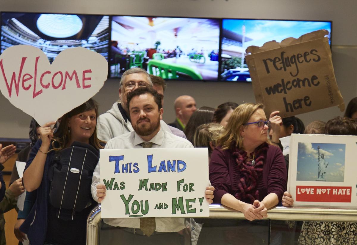 Welcoming community members gather at the airport to welcome a refugee family. People holding different signs that say Welcome, This Land Was Made for You and Me, and Refugees Welcome stand behind an airport guard rail.