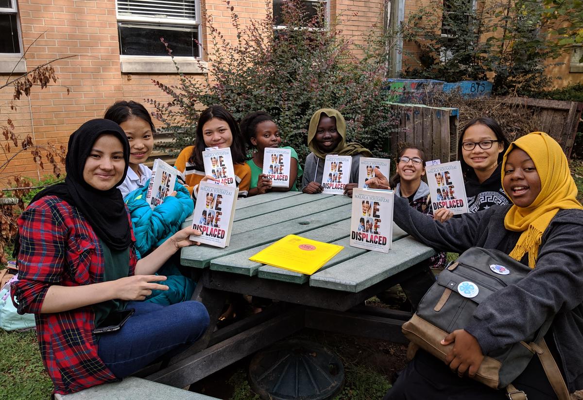 Students read We are Displaced: My Journey and Stories from Refugee Girls Around the World. A diverse group of high school age girls are sitting around an outdoor picnic table and holding copies of Malala Yousafzai’s most recent book.