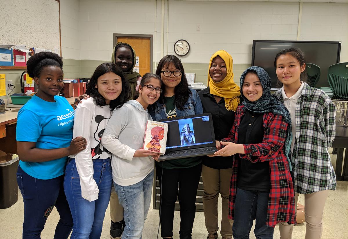 Book Club participants had a surprise Skype session with Jasmine Warga, author of Other Words for Home. A diverse group of female high school students are standing together in a classroom. One student holds a laptop with an open Skype call and author Jasmine Warga is on the screen.