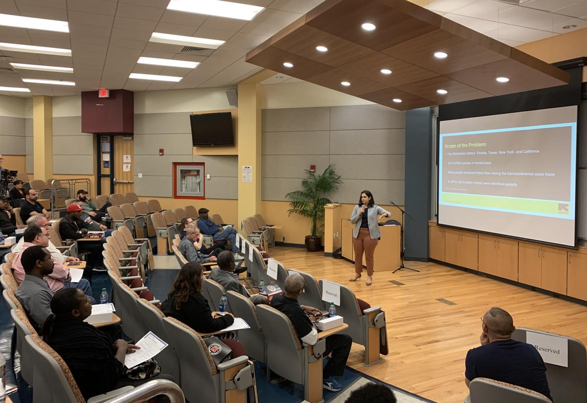 The IRC's Caridad Mas-Batchelor delivers trafficking awareness training to Uber drivers in partnership with Florida's Attorney General, Ashley Moody. A woman stands on a stage in front of several Uber drivers, an IRC presentation is shown on the screen in the background.