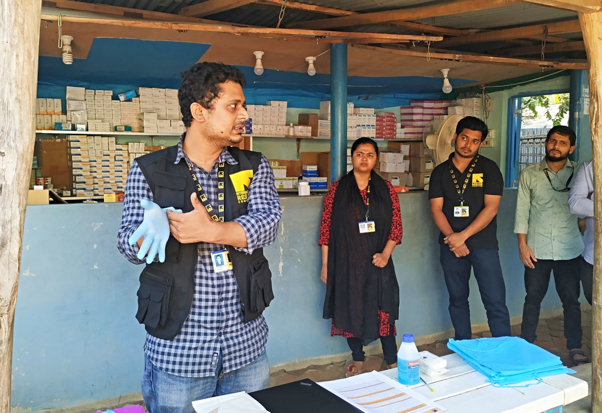 In Coxs Bazar refugee camp in Bangladesh, Dr. Mahmud demonstrates the proper way to put on gloves for health workers and health volunteers. Dr. Mahmud puts on a glove to demonstrate the proper technique, while health workers and health volunteers watch him.