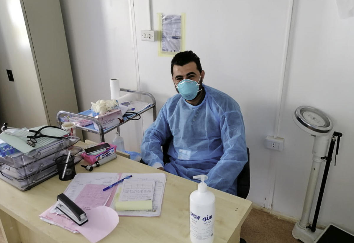 Dr. Hazem at the IRC's clinic. Our health clinics in Jordan, including in Azraq and Za'atari refugee camps, remain open in spite of a national lockdown Dr Hazem sits behind his desk at the IRC's clinis. He is wearing a surgical mask and scrubs.