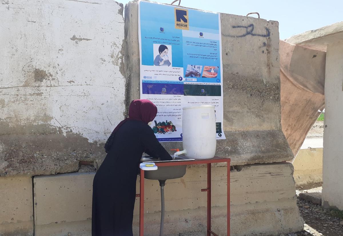 A woman washes her hands at an IRC handwashing station at the main entry gate to the city of Lashkar Gah, in Helmand Province in Afghanistan. A woman washes her hands at an IRC handwashing station with an informational poster on a wall above her head.