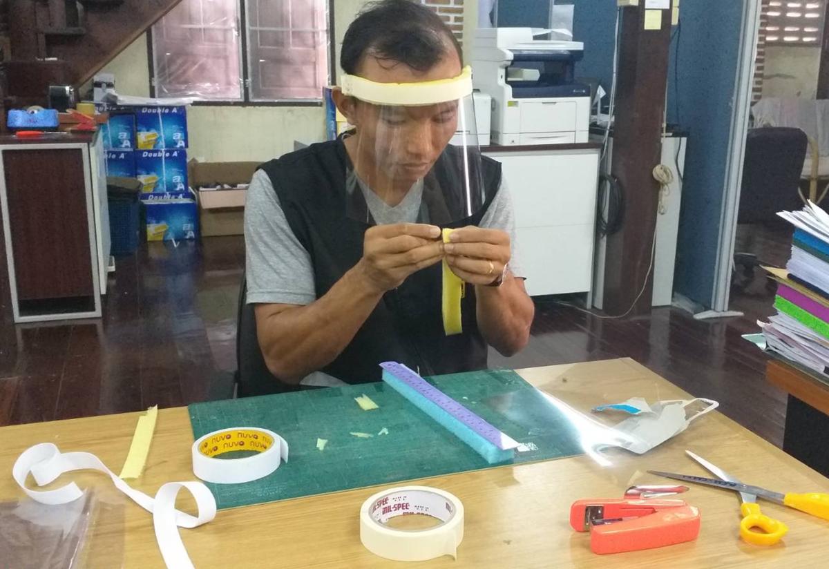 An IRC staff member in a refugee camp in Thailand sits at a desk wearing a face shield. The desk has scissors, a ruler, and tape, and he is using the supplies to create face shields.