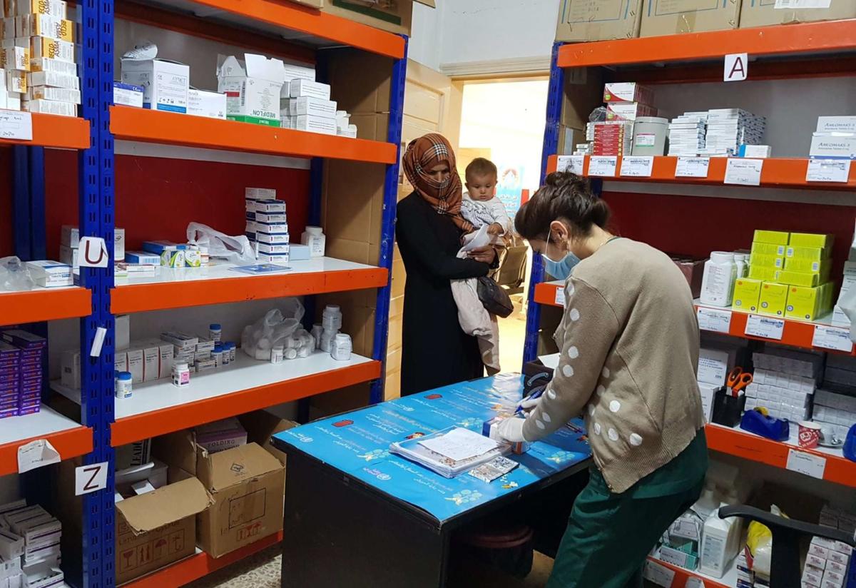 At a pharmacist in NE Syria, a pharmacist wearing a mask and gloves prepares medication while a mother holding a baby waits to get her medicine.