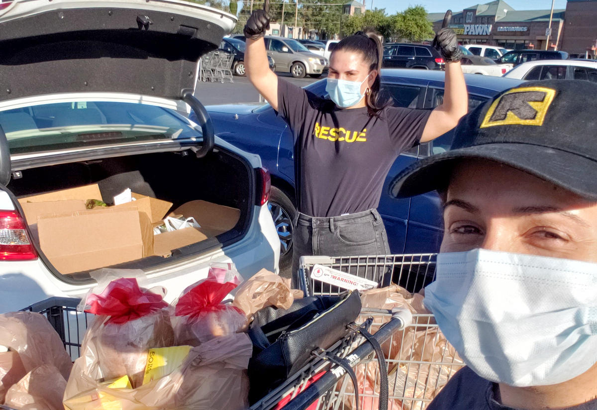 Two IRC staff members wearing IRC shirts and hats stand next to a car with an open trunk. In the trunk are groceries they recently bought for refugee and immigrant families. 