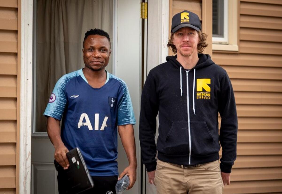 An IRC staff member, prior to strict social distancing, delivering laptops to new refugee arrivals and helping them set up internet in their new homes. An IRC staff member stands next to an IRC client holding a laptop.