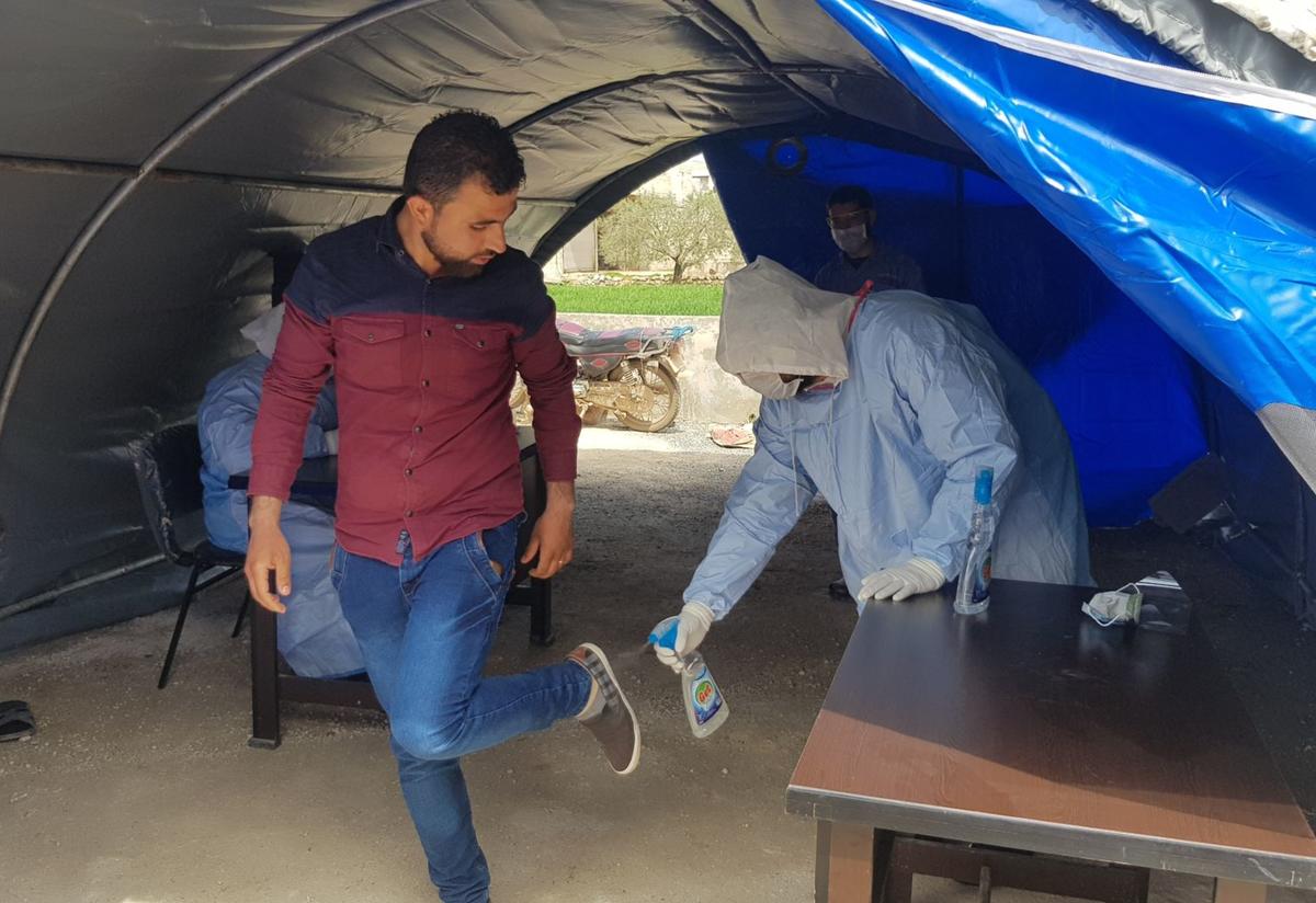 A health worker disinfects a patient’s shoes before he enters a clinic in northwest Syria. Within a tent outside of an IRC health clinic, a health worker in full PPE sprays disinfectent on the bottom of a clients shoes. Another health worker in full PPE sits at a desk behind them.