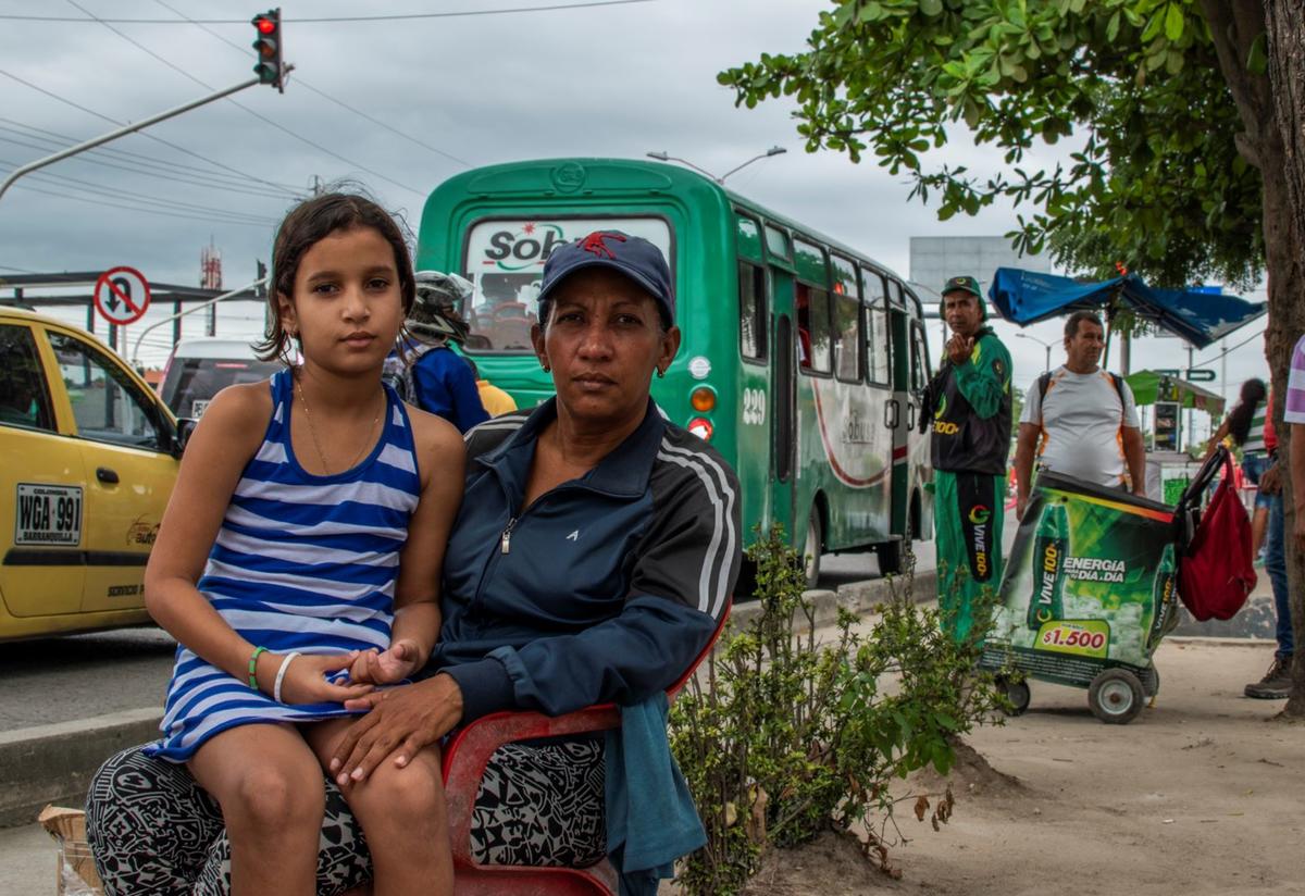 Venezuelan mother Karina and her ten-year-old daughter Geicelis, in a photo taken before the coronavirus pandemic. Venezuelan mother Karina sits with her ten-year-old daughter Geicelis on her lap. They are sitting outside looking at the camera. There is a green bus behind them.