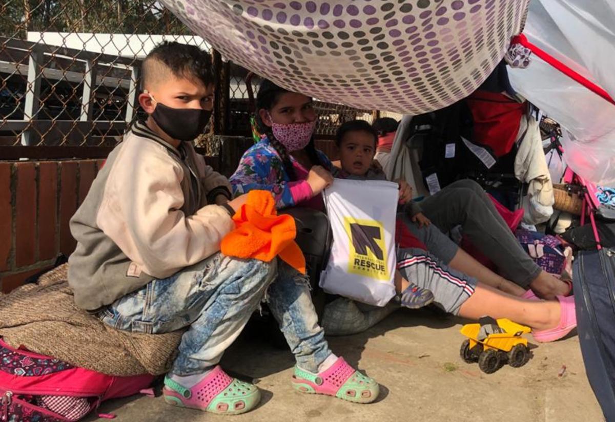 A mother with two kids sit under a makeshift tent surrounded by their luggage. They are all wearing masks and are looking directly at the camera. 
