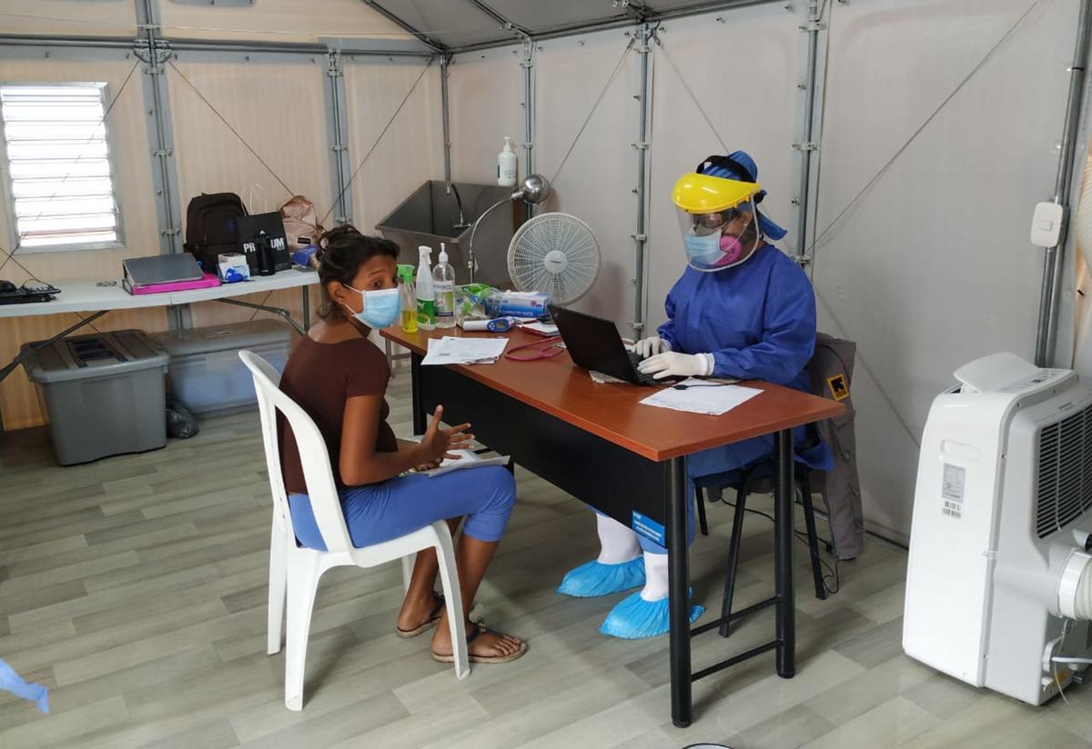 A woman sits at a chair at a table with doctor in full PPE opposite her. They are in a temporary clinic set up by the IRC at the Simón Bolívar bridge. 
