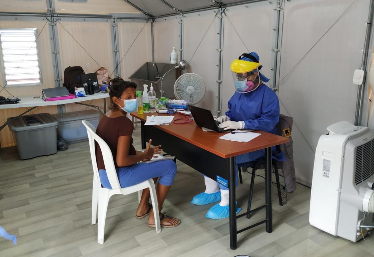 A woman receives medical care from the IRC at the Simón Bolívar bridge. A woman sits at a chair at a table with doctor in full PPE opposite her. They are in a temporary clinic set up by the IRC at the Simón Bolívar bridge.