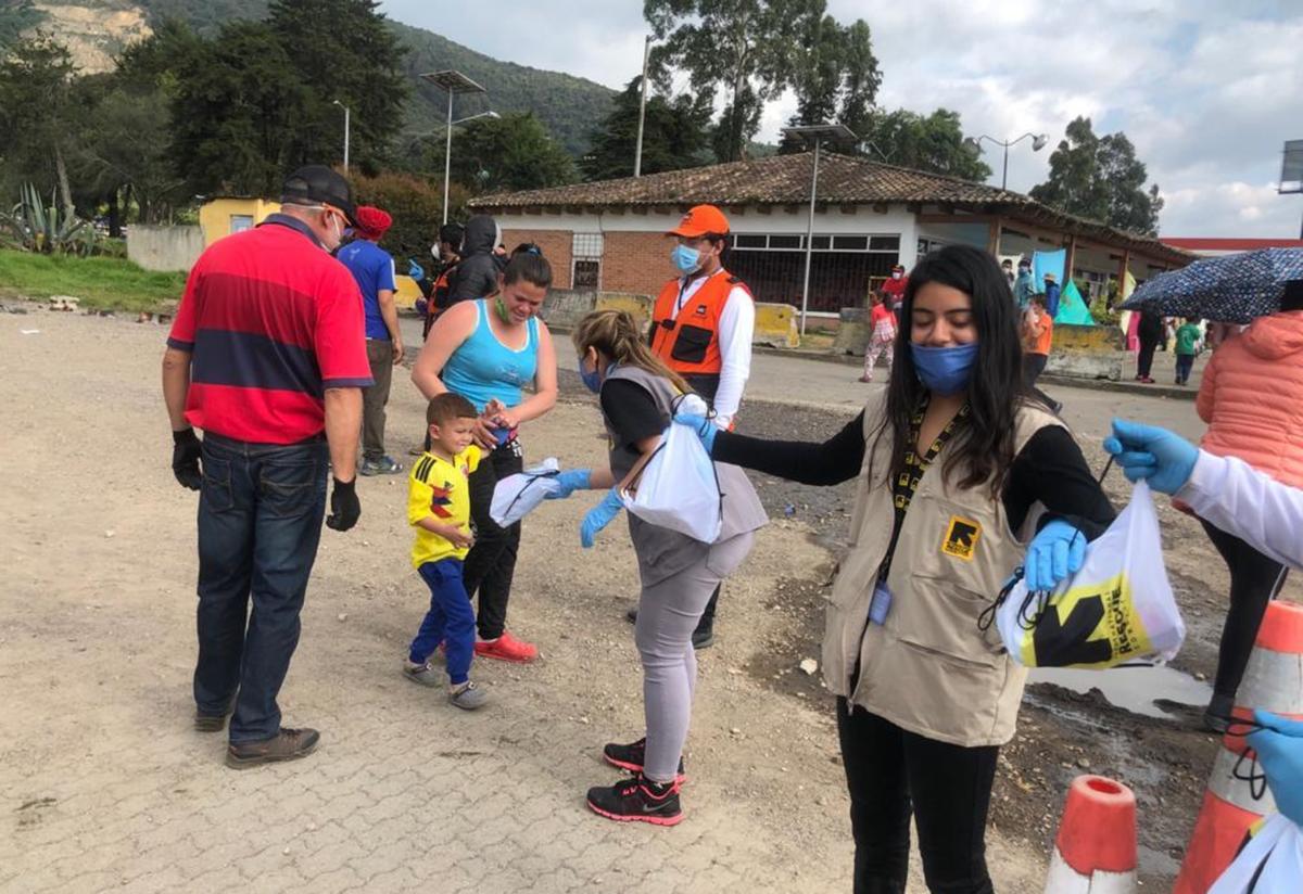 IRC teams deliver hygiene kits, drinking water, snacks and hot food to Venezuelan migrants walking to the border. Two IRC staff members stand in a line passing out supplies outside near the Bogota-Chia highway. A family, including a young boy, are given supplies.