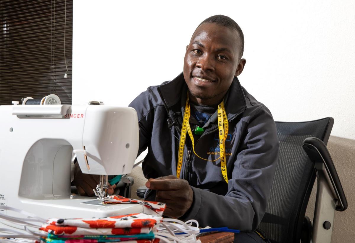 Jonathan Amissa and the sewing machine he uses to create masks. Jonathan Amissa sits at a desk with his sewing machine. He has a yellow tape measure around his neck and there is a stack of red masks on the desk.