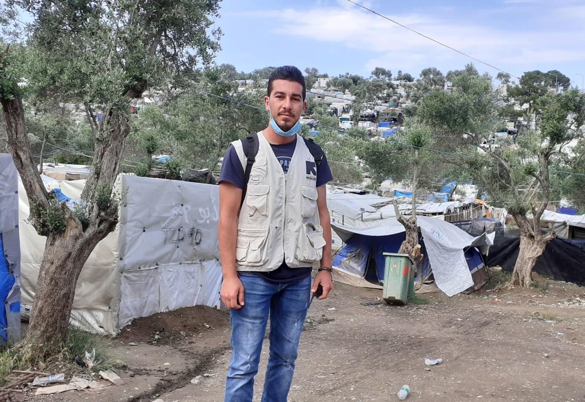 Shadi Shadi stands in front of a tree and a tent in a refugee camp. He is wearing a mask around his neck and an IRC vest.
