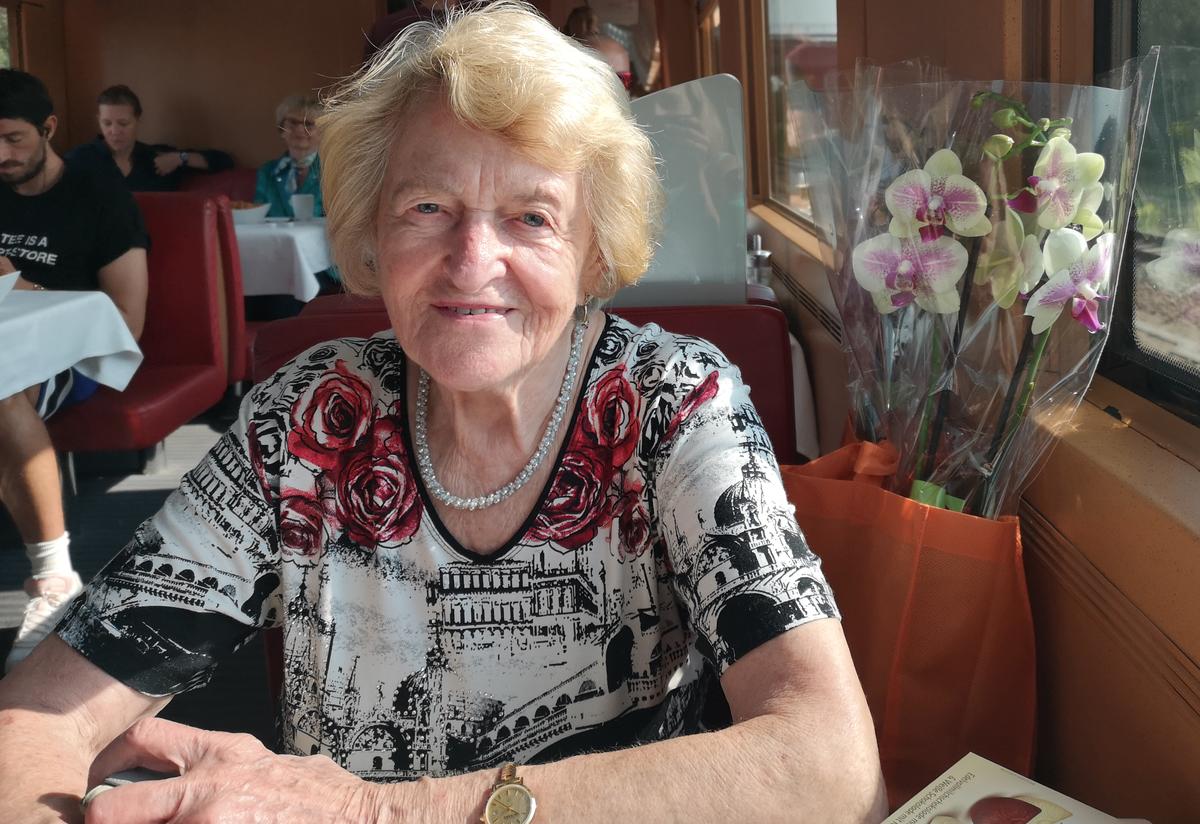 Irmgard Irmgard sits at a table at a restaurant. She is wearing black and white shirt with red roses. There is a bouquet of flowers behind her.