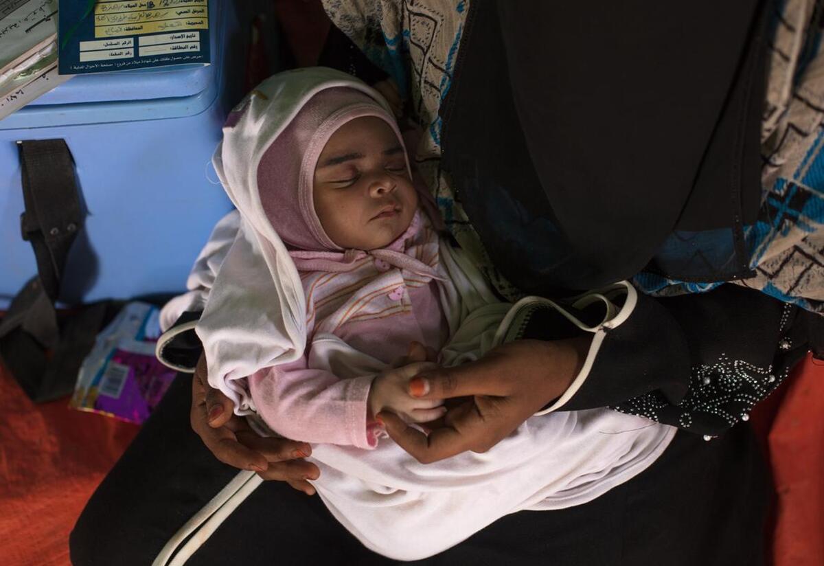 Baby Enkath, which means rescue, sleeps on her mothers lap. Wearing a pink onesie, baby Enkath sleeps on her mothers lap. They are inside a tent.