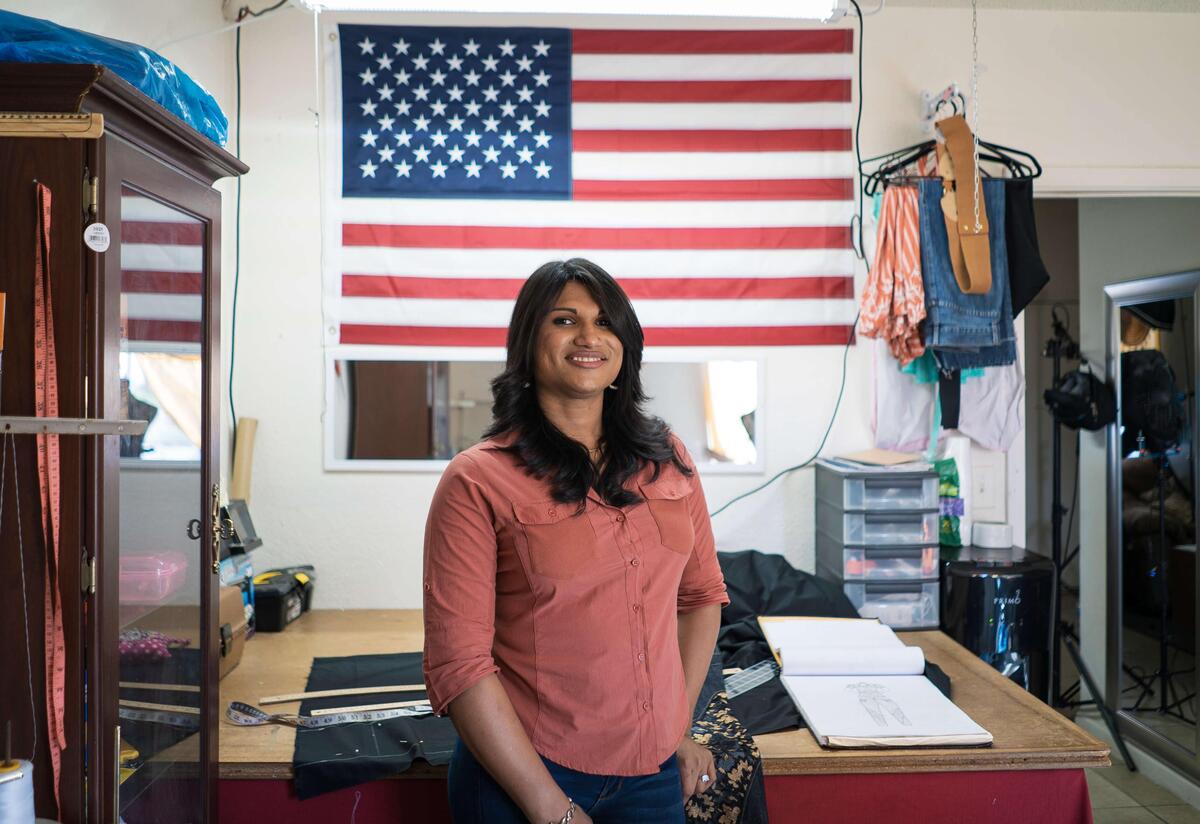 Lincy Sopall stands in her studio for her fashion design business. She is wearing a pink button down shirt and smiling. Behind her is a desk, supplies for her work, and a large American flag. 