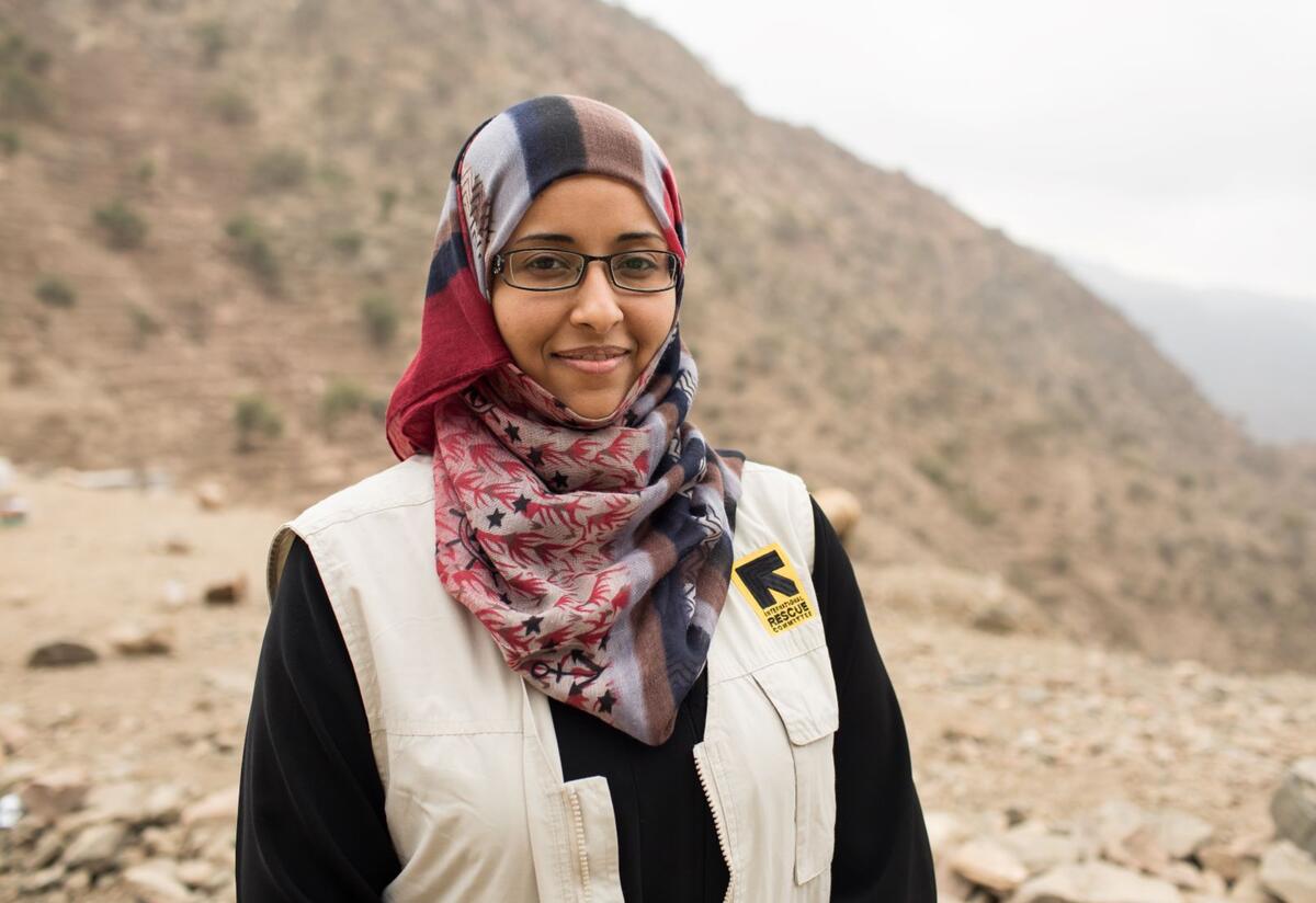 Dr. Rasha Rashed stands outside in front of a mountain in Yemen. She is wearing a colorful headscarf and an IRC vest. 