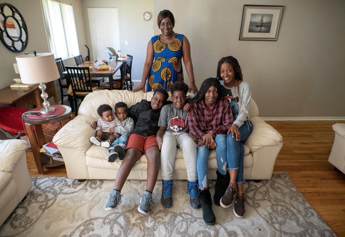 Christelle, her mother Jacqueline Uwumeremyi, and their family Jacqueline Uwumeremyi stands behind a couch where all of her children sit. There are 6 children, 4 boys and 2 girls.