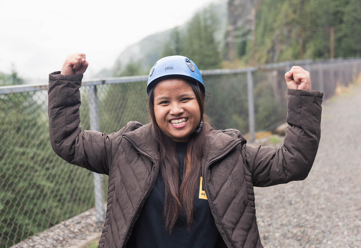Close-up photo of smiling teenage girl with arms up in a strong pose. Girl wears winter coat and rock climbing helmet, and is outdoors in wooded area.