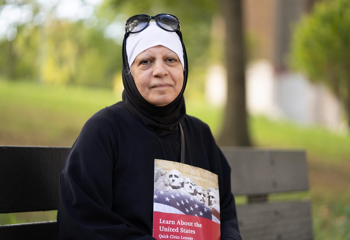Maha sits on a bench in Astoria Park looking at the camera. She is holding a citizenship exam study book. 